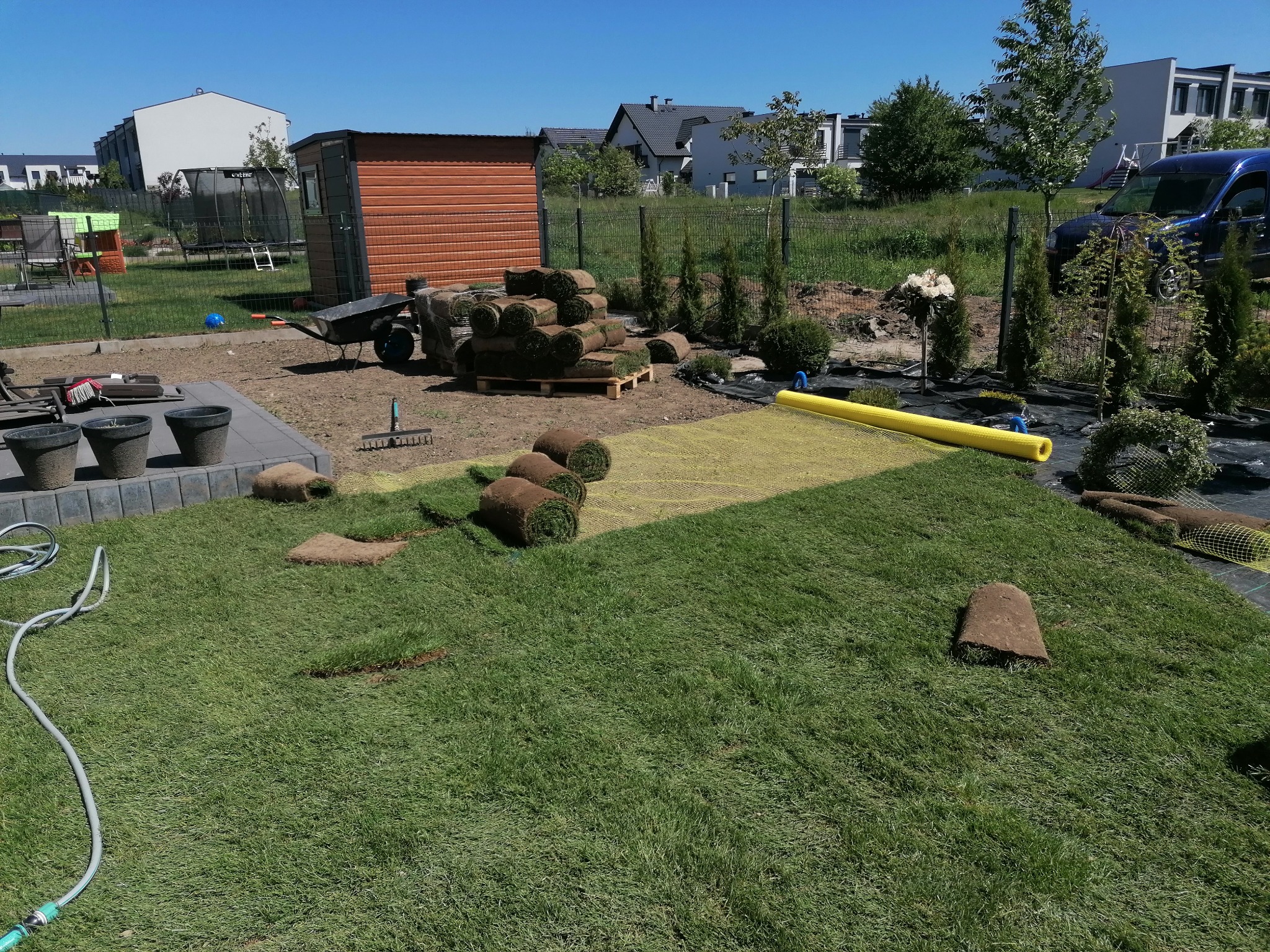 Rozłożona trawa z rolki na działce, z widocznymi rolkami trawy na palecie, żółta siatka wzmacniająca podłoże, oraz fragmenty trawy przygotowane do ułożenia, a w tle zabudowa jednorodzinna...