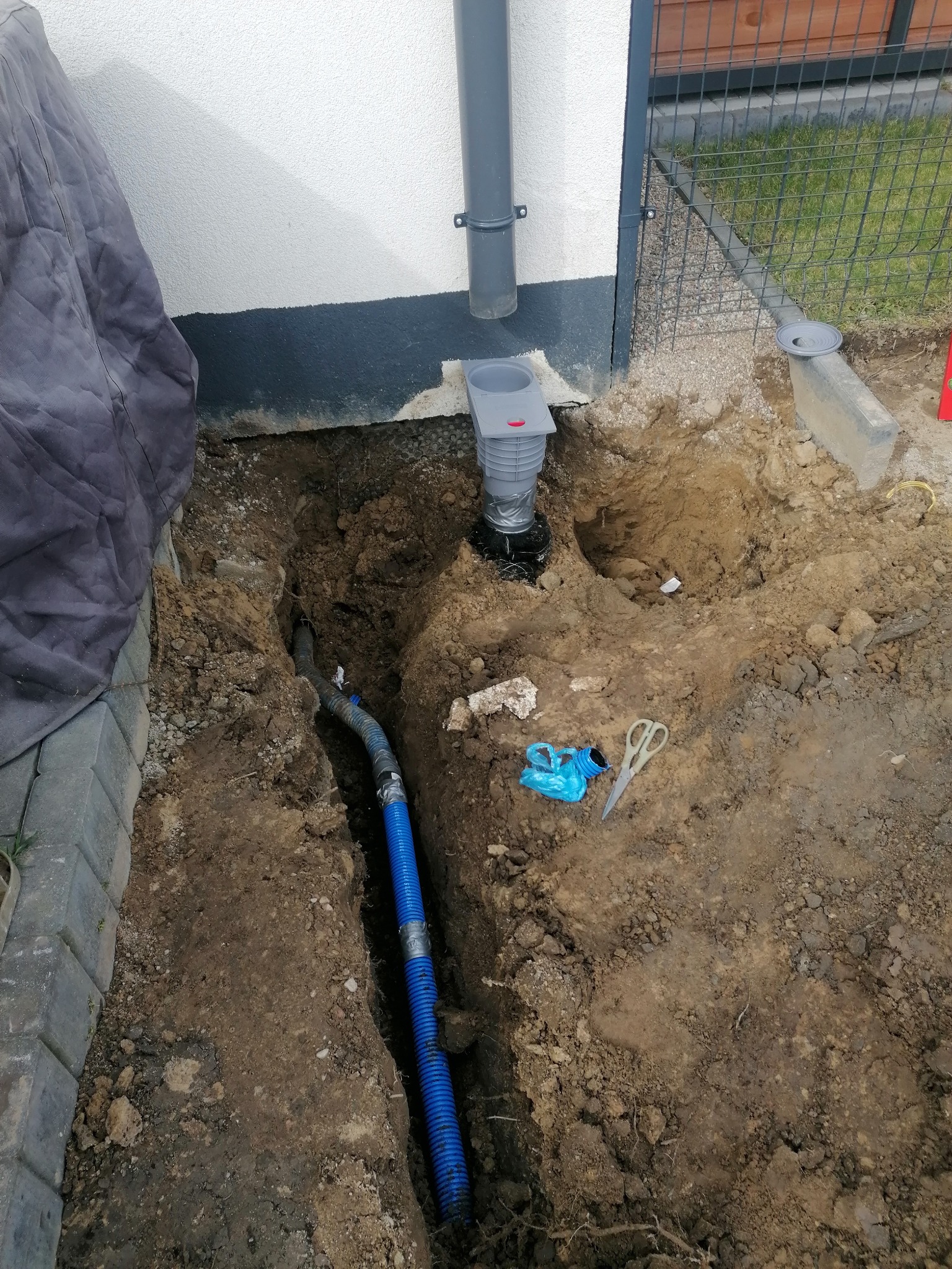 Instalacja systemu odprowadzania wody deszczowej: wykop z niebieską rurą drenażową, studzienka rewizyjna z szarą kratką, narzędzia (nożyczki, worek) leżące na ziemi, widok z góry.