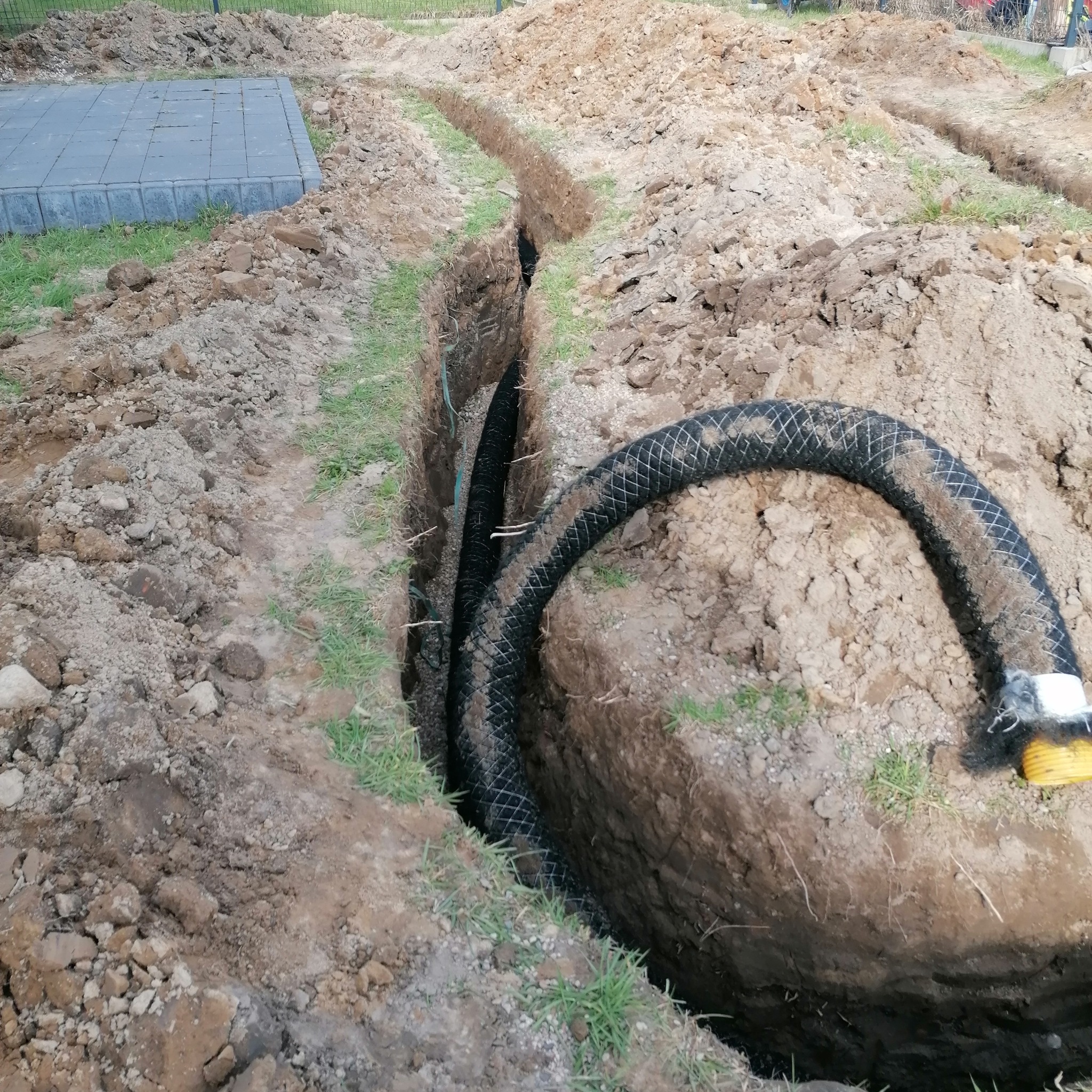 Wykop z rurą drenażową w ziemi, widoczny fragment trawnika i kostka brukowa w tle.