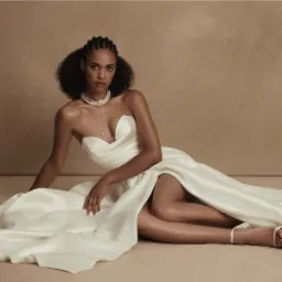 Modelka w białej, satynowej sukni bez ramiączek z wysokim rozcięciem, leżąca na podłodze w studio, z perłowym naszyjnikiem i delikatnymi sandałkami.