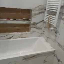 Biała wanna obudowana płytkami imitującymi marmur, drewniane wnęki w ścianie nad wanną, grzejnik drabinkowy na ścianie wyłożonej marmurowymi płytkami.