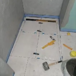 Szare płytki podłogowe w trakcie układania w łazience, widoczne krzyżyki dystansowe, poziomica, gumowy młotek i wiadro z zaprawą.