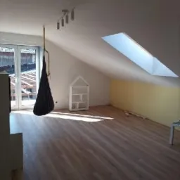 Poddasze z oknem dachowym, drewnianą podłogą, pomalowaną na żółto ścianą i wiszącym czarnym hamakiem, widoczne białe drzwi balkonowe i biały regał w kształcie domku.