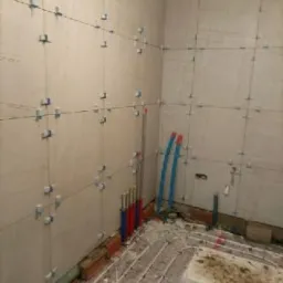 Ściana w trakcie układania płytek z użyciem krzyżyków dystansowych, widoczne rury instalacji ogrzewania podłogowego i piony instalacyjne.
