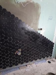 Układanie czarnych, heksagonalnych płytek na ścianie w łazience, widoczne fragmenty surowej ściany i instalacji.