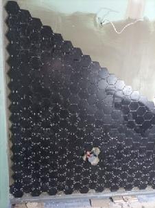 Ściana pokryta czarnymi, heksagonalnymi płytkami w trakcie układania, widoczne niedokończone fragmenty i przewody elektryczne.