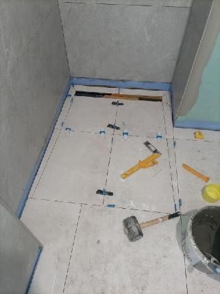 Szare płytki podłogowe w trakcie układania w łazience, widoczne krzyżyki dystansowe, poziomica, gumowy młotek i wiadro z zaprawą.