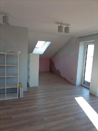 Poddasze po remoncie: świeżo położona podłoga z paneli, ściana częściowo przygotowana do malowania z widocznymi fragmentami gładzi, okno dachowe i białe regały.