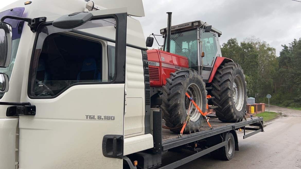 Czerwony traktor Massey Ferguson 3680 zabezpieczony pomarańczowymi pasami transportowymi na platformie białej ciężarówki MAN TGL 8.180, widok z boku na tle drzew.