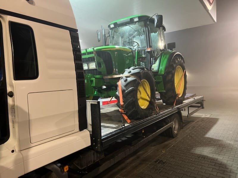 Zielony traktor John Deere zabezpieczony pomarańczowymi pasami na platformie lawety, widoczny fragment białej kabiny samochodu transportowego, oświetlenie w tle sugeruje transport nocą lub...