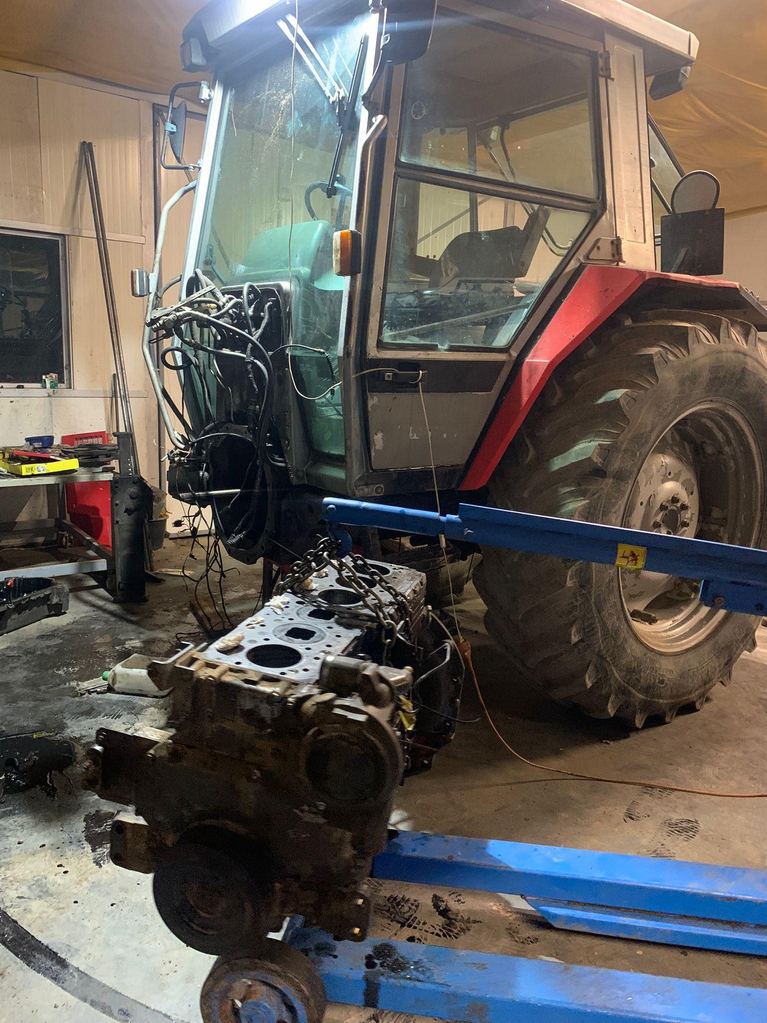 Częściowo rozebrany traktor z wyjętym silnikiem na niebieskim wózku warsztatowym, widoczne przewody i elementy mechaniczne.