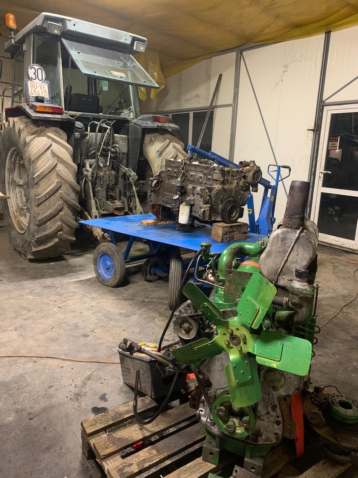 Warsztatowa scena z demontażu silnika ciągnika: odłączony silnik na wózku warsztatowym z podnośnikiem, drugi silnik z zielonym wentylatorem na palecie, w tle ciągnik z otwartą kabiną.
