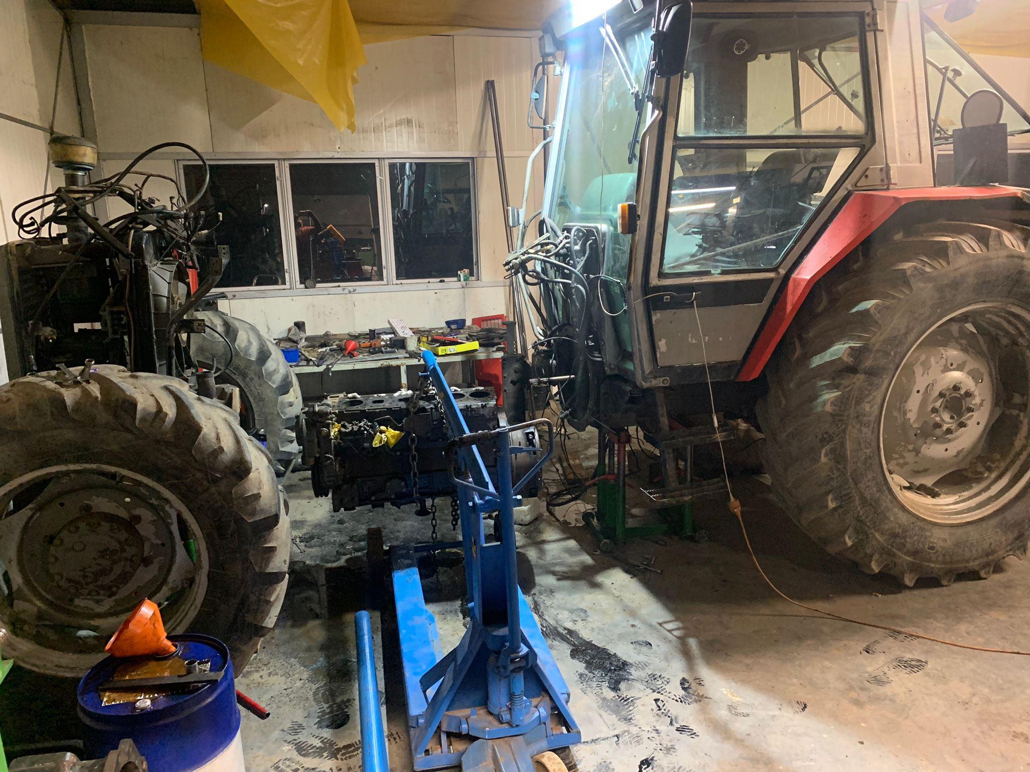 Częściowo zdemontowany traktor w warsztacie, silnik na niebieskim wózku warsztatowym, widoczne narzędzia i akcesoria.