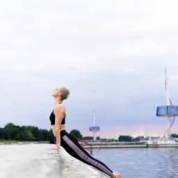 Kobieta z krótkimi blond włosami w czarnym sportowym topie i legginsach wykonuje pozycję jogi na betonowym nabrzeżu, z widokiem na wodę i konstrukcje w tle.