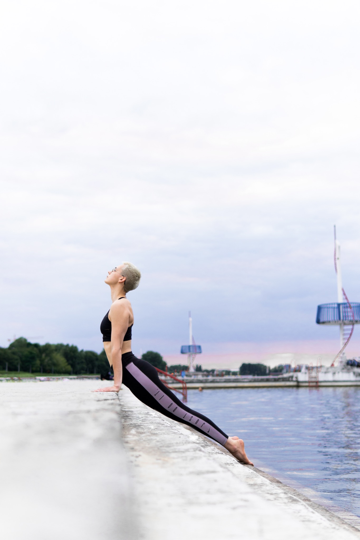 Kobieta z krótkimi blond włosami w czarnym sportowym topie i legginsach wykonuje pozycję jogi na betonowym nabrzeżu, z widokiem na wodę i konstrukcje w tle.