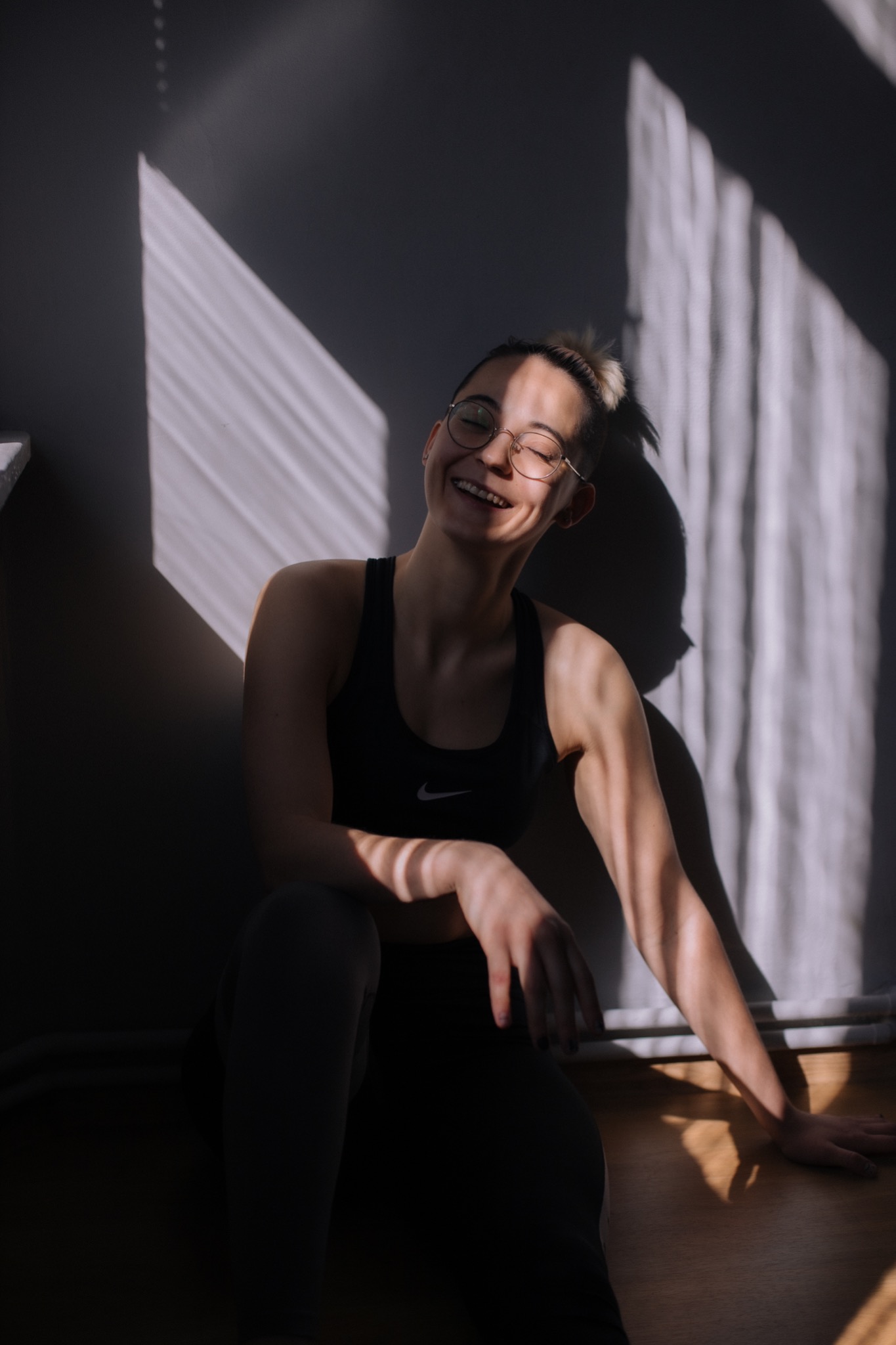 Uśmiechnięta kobieta w okularach siedzi na podłodze w sportowym stroju, oświetlona promieniami słońca padającymi przez okno.