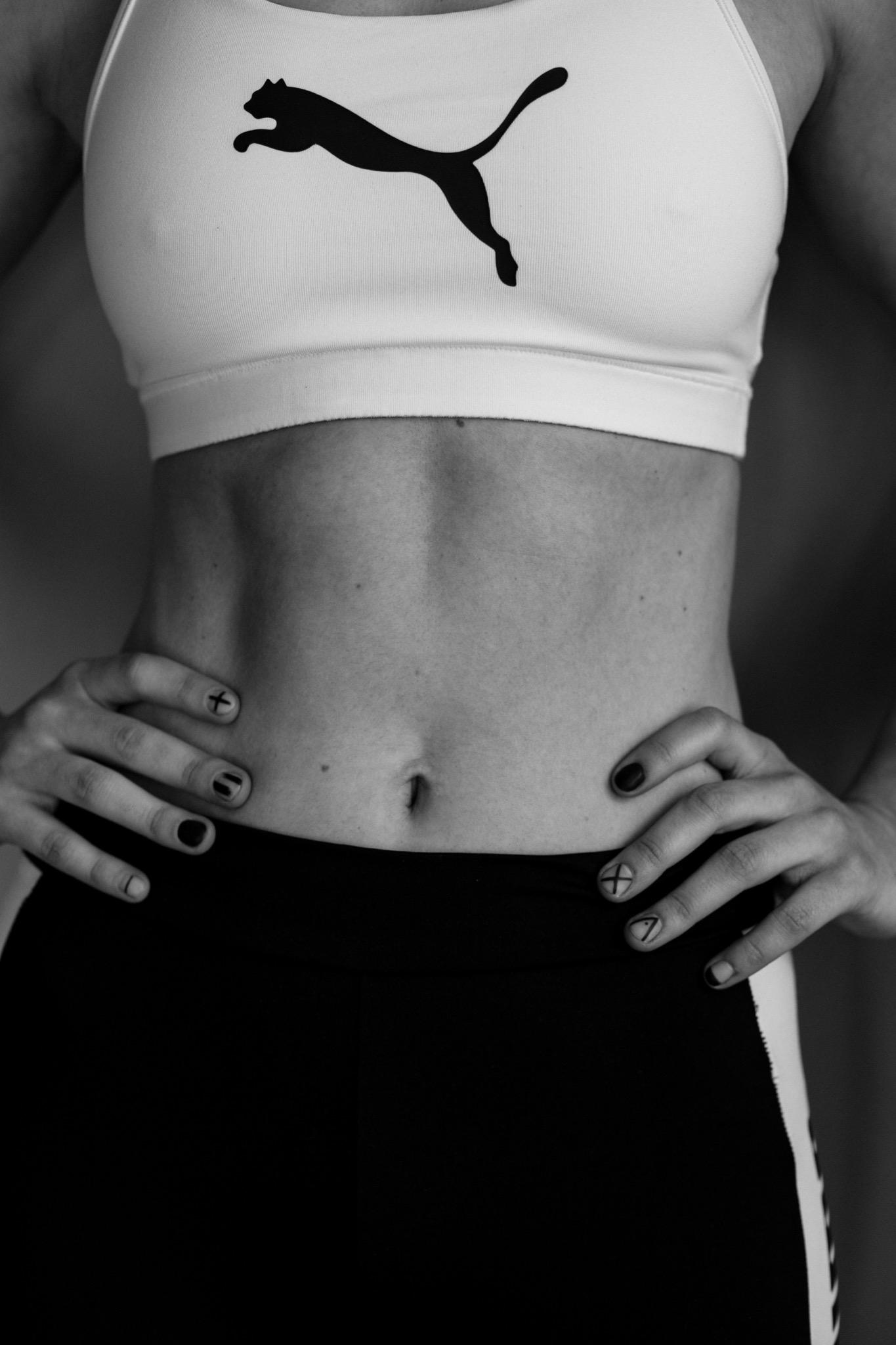 Czarno-białe zdjęcie częściowo widocznej kobiety w sportowym topie z logo Puma, z rękami na biodrach i pomalowanymi paznokciami w geometryczne wzory, ukazujące wyrzeźbiony brzuch.