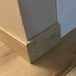 Zamknięcie narożnika listwą przypodłogową w Łodzi, dopasowaną kolorystycznie do paneli podłogowych, przy białej futrynie drzwi.