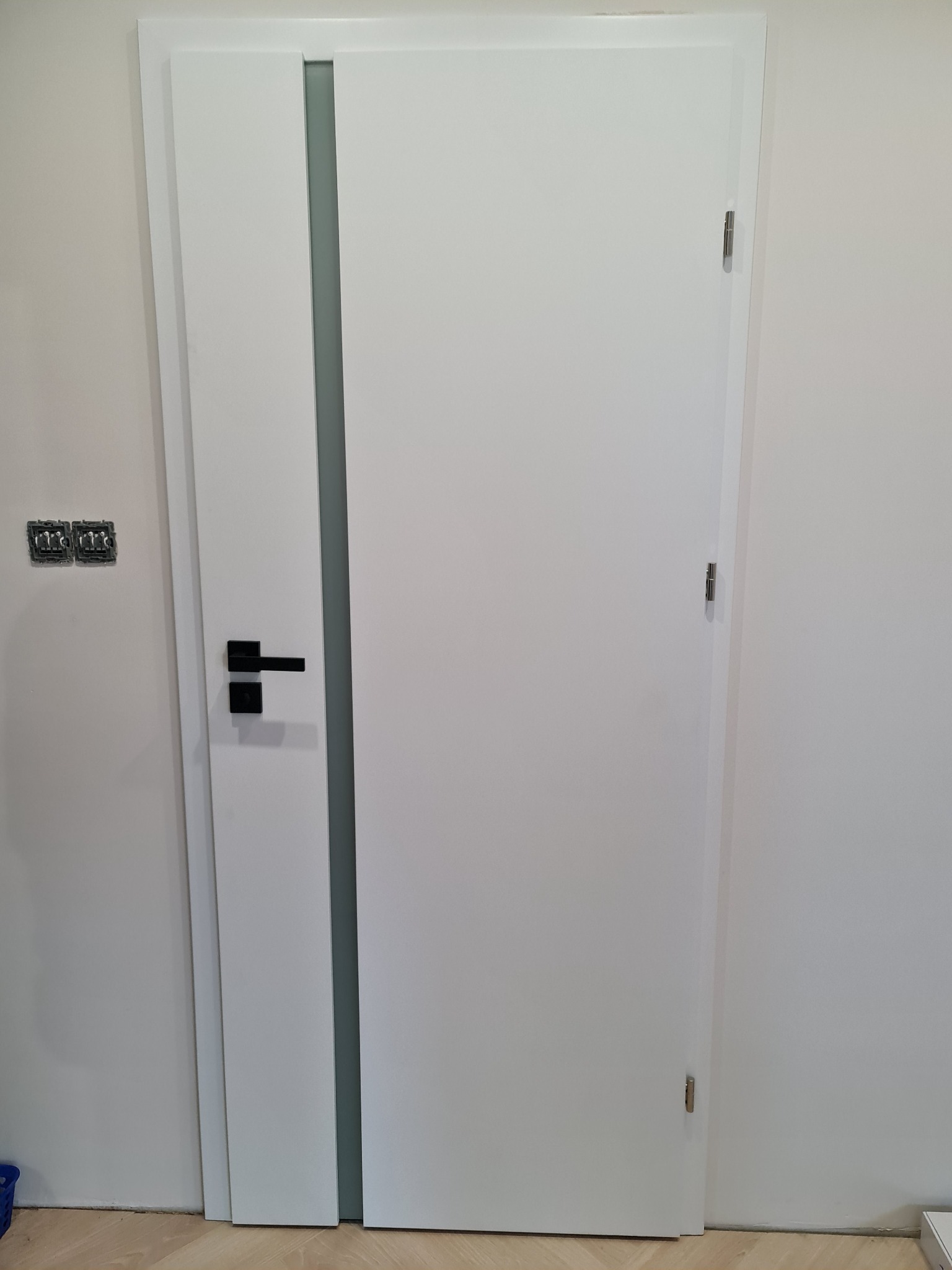 Białe drzwi wewnętrzne z czarną klamką i szarym pionowym paskiem, zamontowane w jasnym pomieszczeniu, widoczne gniazdka elektryczne po lewej stronie.