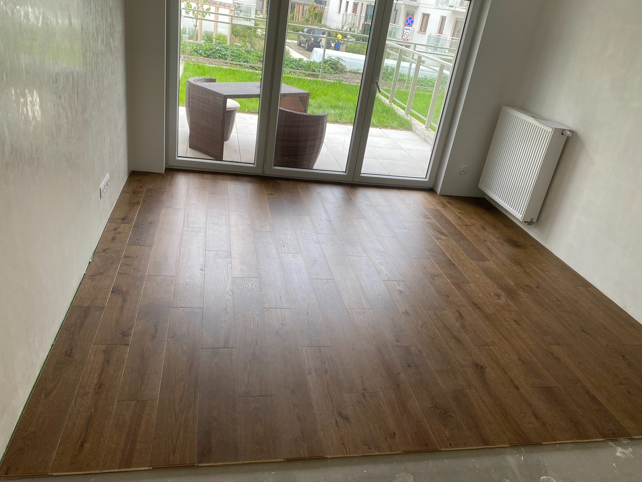 Nowo ułożona podłoga z ciemnych paneli w jasnym pomieszczeniu z widokiem na taras z meblami ogrodowymi.