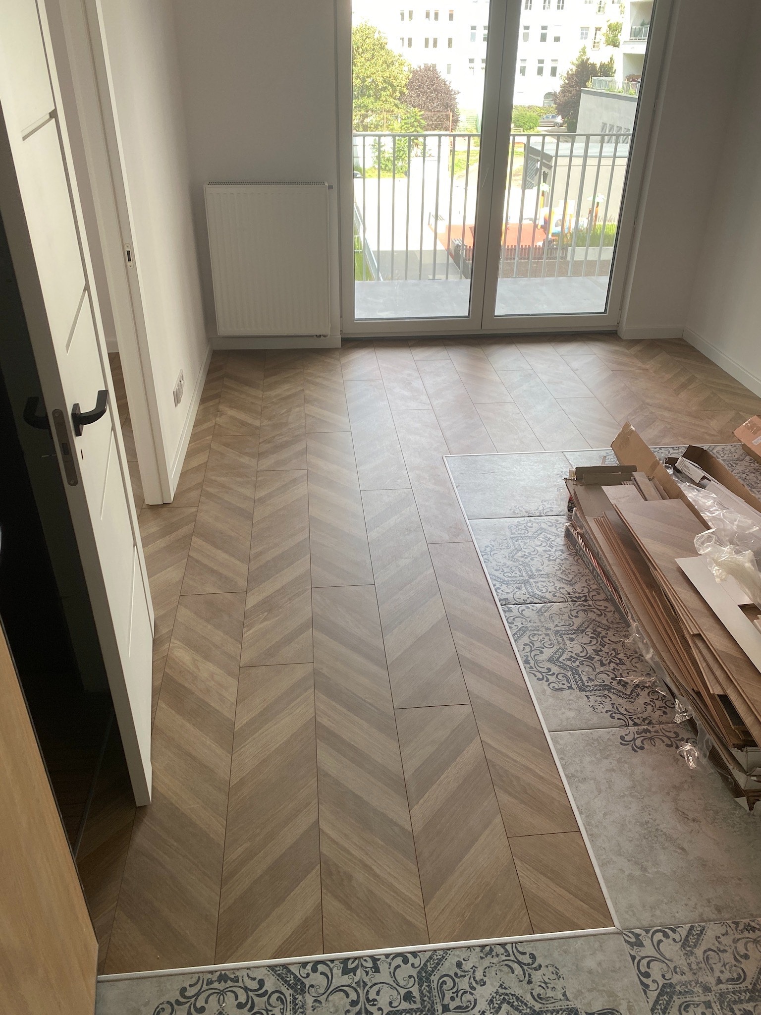 Ułożona podłoga z paneli w jodełkę, widoczne paczki paneli obok, płytki dekoracyjne na podłodze, drzwi balkonowe i grzejnik w tle.