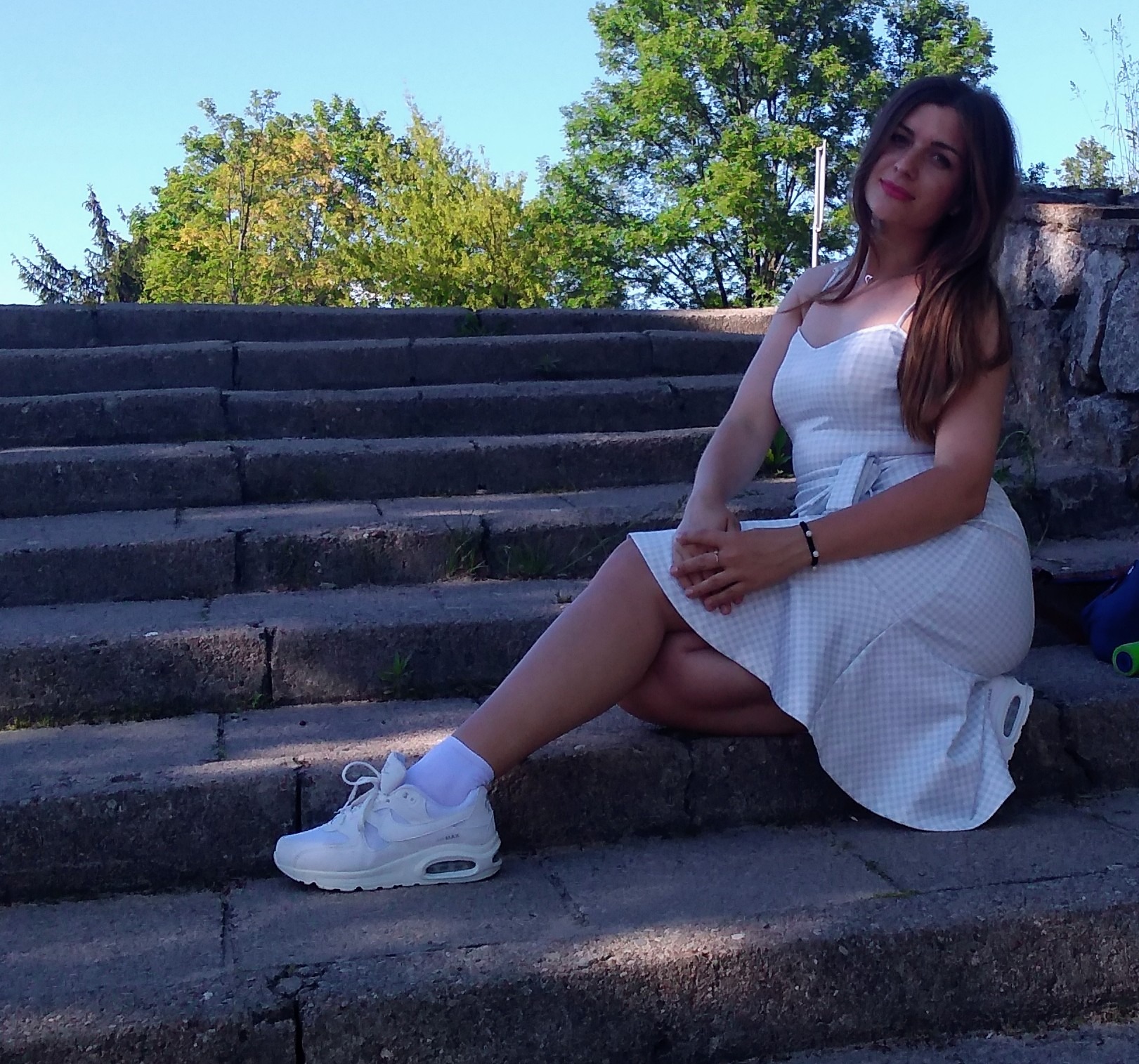 Kobieta w letniej sukience siedzi na kamiennych schodach w parku.