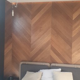 Zakład Malarski Tomasz Frasek - Ściana z drewnianych paneli ułożonych w jodełkę, widoczne niedokończone podłączenie lampy sufitowej i fragment sypialni z łóżkiem.