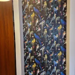 Fragment ściany z tapetą w ciemnych kolorach, pokrytą wzorem stylizowanych, kolorowych ptaków i kwiatów, widoczne krawędzie ściany i fragment drewnianych drzwi po lewej stronie.