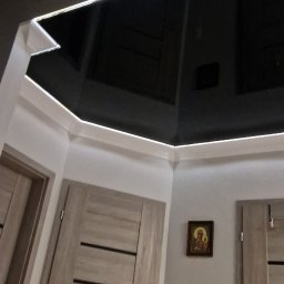 Zakład Malarski Tomasz Frasek - Nowoczesne wykończenie wnętrza: czarny, błyszczący sufit z oświetleniem LED, szare ściany z obrazem i drewniane drzwi z ciemnymi paskami, widoczne pod kątem.