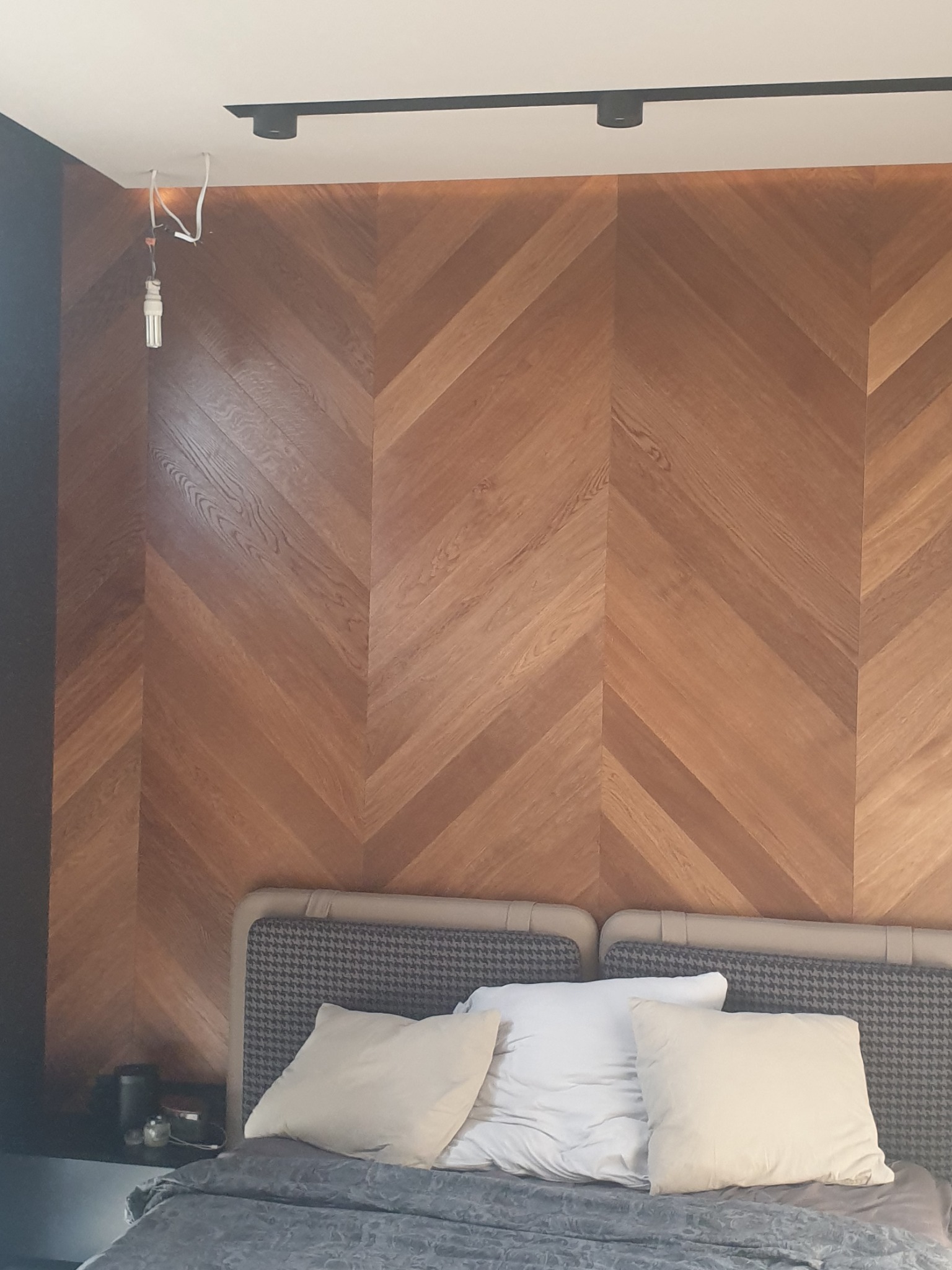 Ściana z drewnianych paneli ułożonych w jodełkę, widoczne niedokończone podłączenie lampy sufitowej i fragment sypialni z łóżkiem.