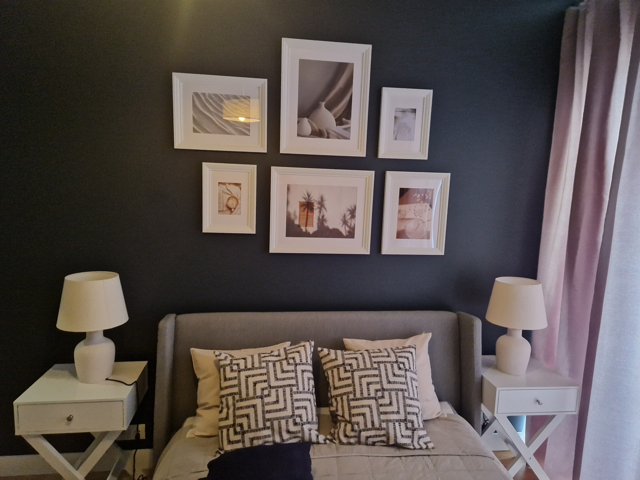 Aranżacja sypialni z ciemną ścianą, galerią sześciu obrazków w białych ramkach, szarą tapicerowaną ramą łóżka, białymi szafkami nocnymi z lampkami oraz zasłonami w odcieniu pudrowego różu.