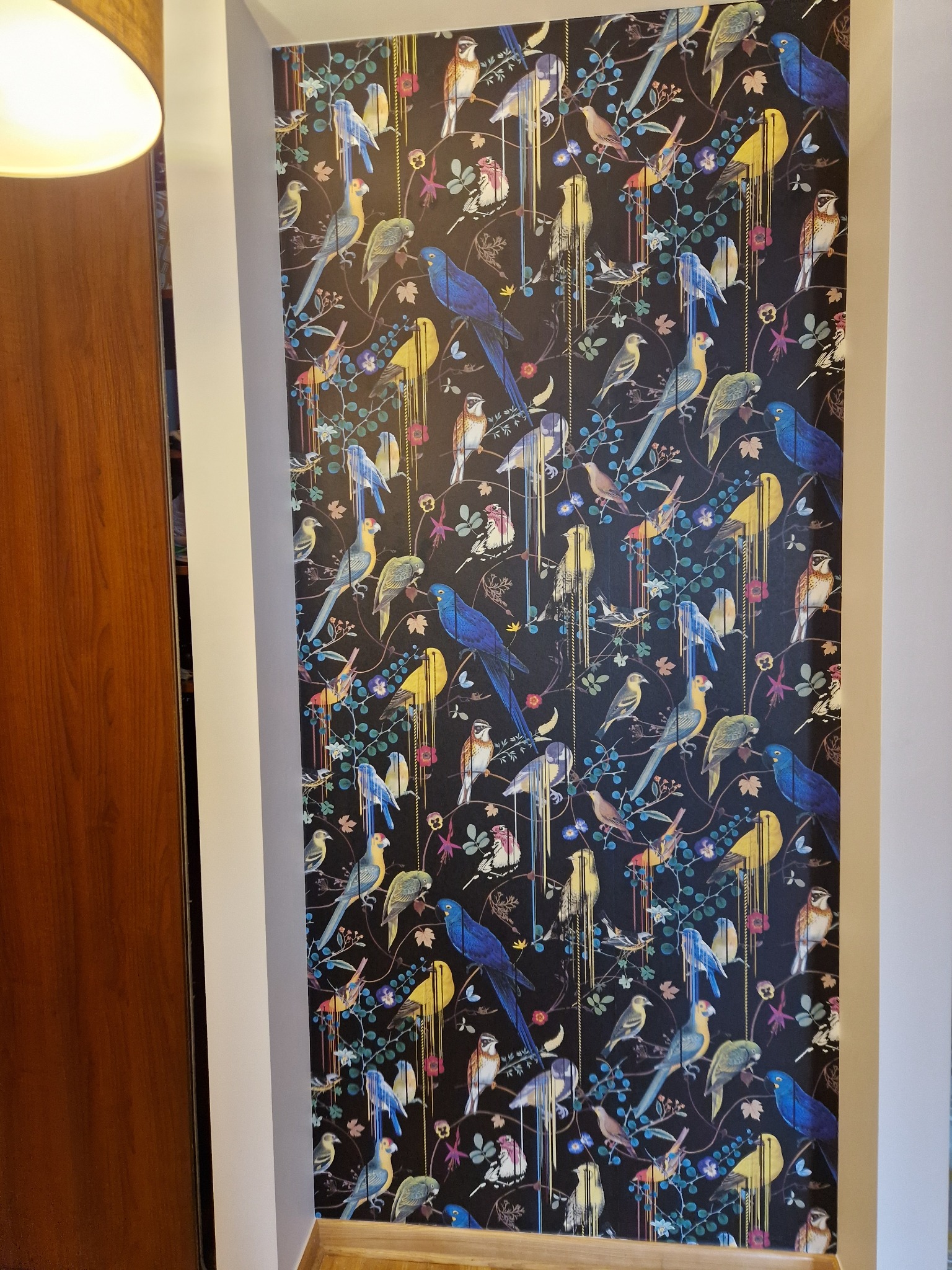Fragment ściany z tapetą w ciemnych kolorach, pokrytą wzorem stylizowanych, kolorowych ptaków i kwiatów, widoczne krawędzie ściany i fragment drewnianych drzwi po lewej stronie.