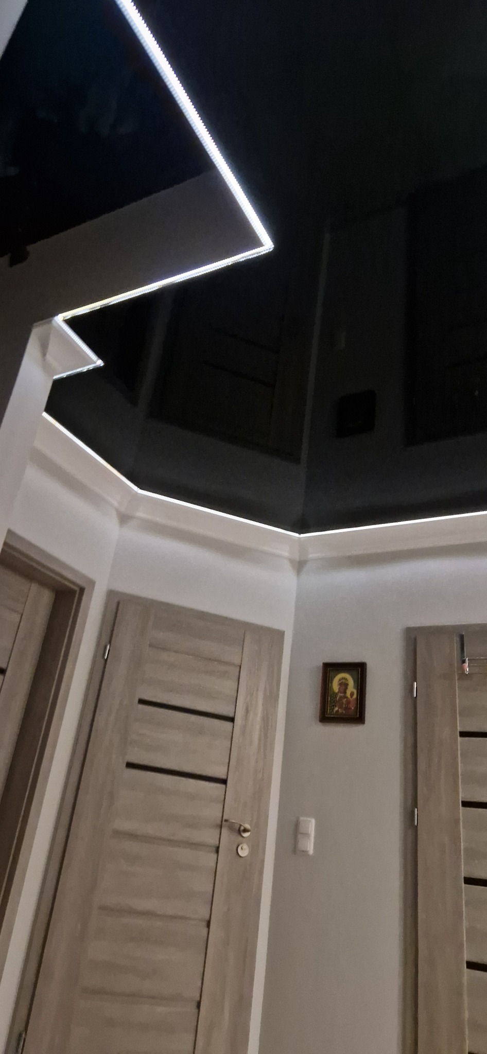 Nowoczesne wykończenie wnętrza: czarny, błyszczący sufit z oświetleniem LED, szare ściany z obrazem i drewniane drzwi z ciemnymi paskami, widoczne pod kątem.