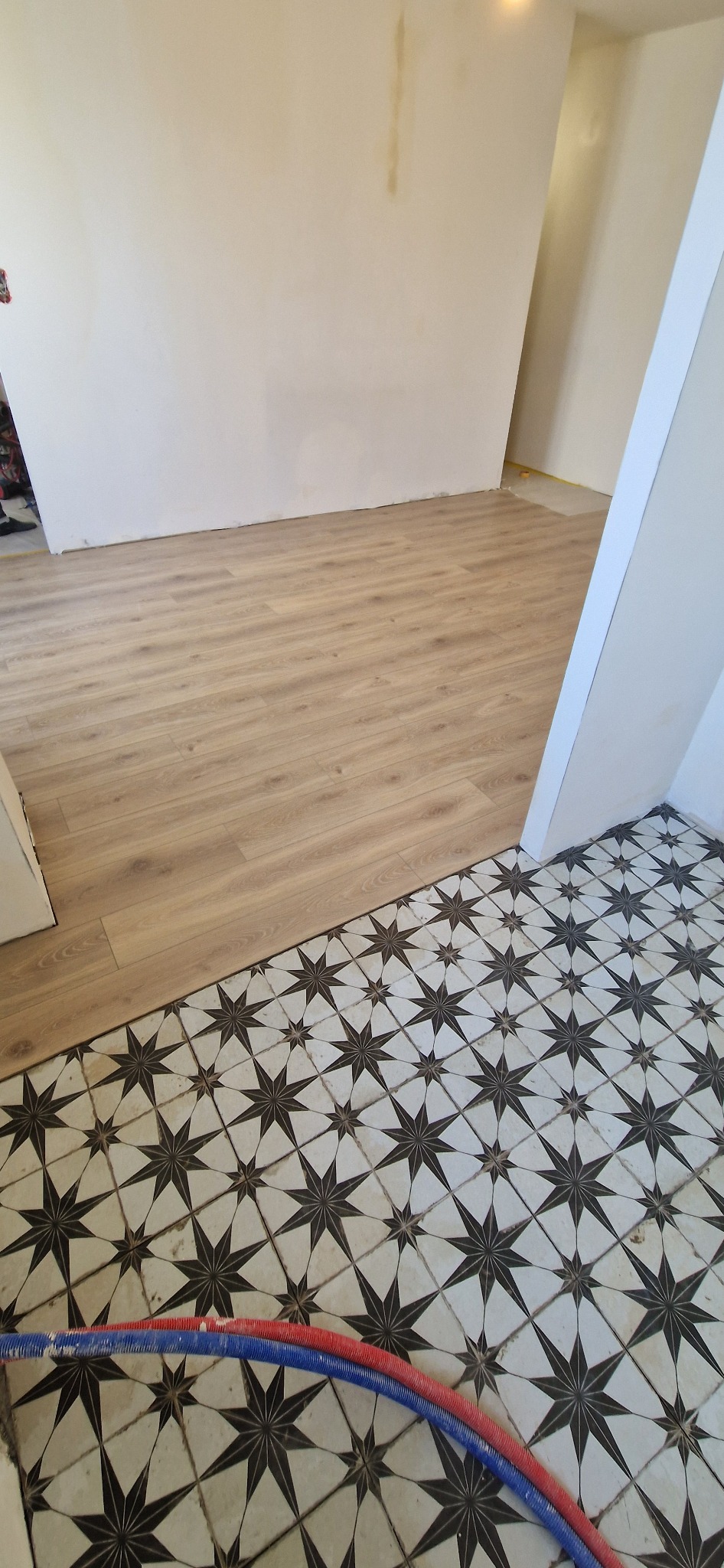 Połączenie dwóch rodzajów podłóg: jasne panele podłogowe i płytki podłogowe w biało-czarne gwiazdy. Widoczne rury instalacyjne w kolorze czerwonym i niebieskim.