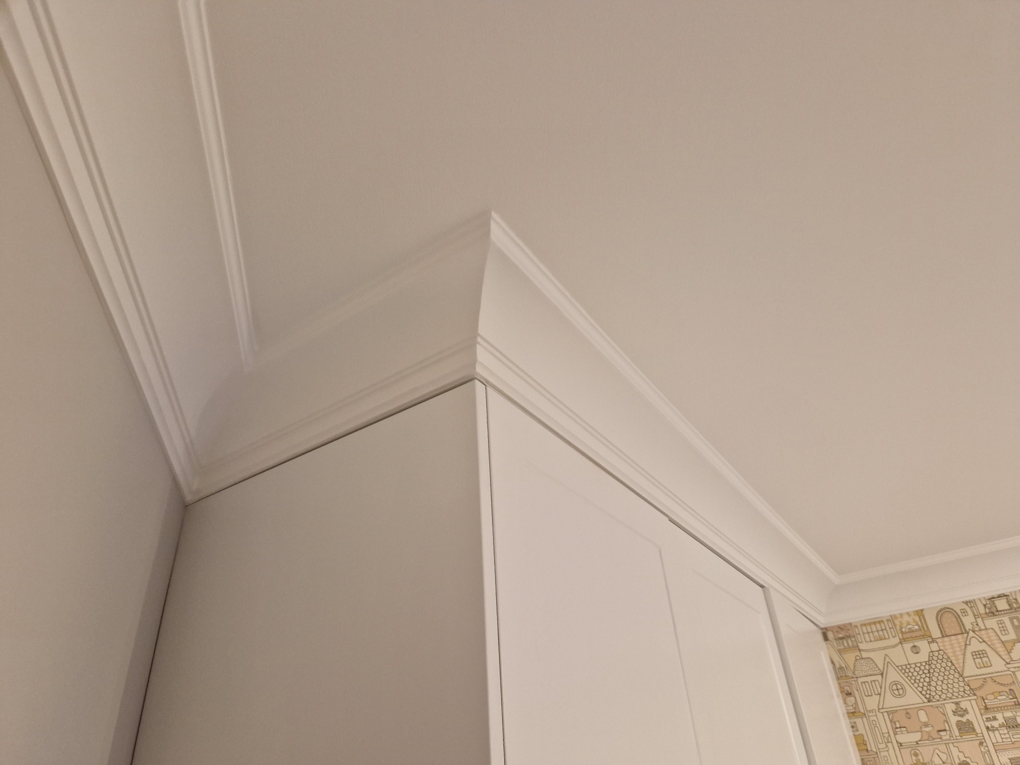 Biała szafa pod sufit, z ozdobną listwą przysufitową, widok z dołu, w tle tapeta w dziecięce wzory.