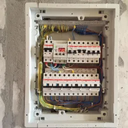 Rozdzielnica elektryczna w białej obudowie zamontowana na surowej, otynkowanej ścianie. Widoczne liczne bezpieczniki, przewody w różnych kolorach oraz wyświetlacz z wartością 230.