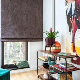Brązowa roleta rzymska z delikatną fakturą w oknie, obok regał z dekoracjami i obraz na ścianie
