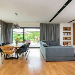 Nowoczesny salon z dużymi oknami, szarymi zasłonami, drewnianym stołem i parkietem, widok na ogród z parasolem.