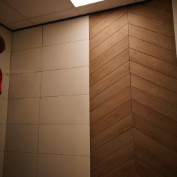 AMAS Andrzej Strzelec - Ściana wykończona jasnymi, kwadratowymi płytkami ceramicznymi po lewej stronie i drewnianą okładziną ułożoną w jodełkę po prawej, oddzielone niebieską taśmą malarską, widoczne oświetlenie sufitowe.
