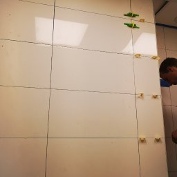 AMAS Andrzej Strzelec - Układanie jasnych, błyszczących płytek ściennych z użyciem plastikowych krzyżyków dystansowych, widoczny fragment twarzy pracownika.