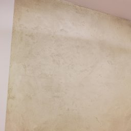 AMAS Andrzej Strzelec - Ściana z efektem betonu architektonicznego w odcieniach beżu i szarości, widoczne nieregularne wzory i faktura, narożnik ściany pomalowany na biało.
