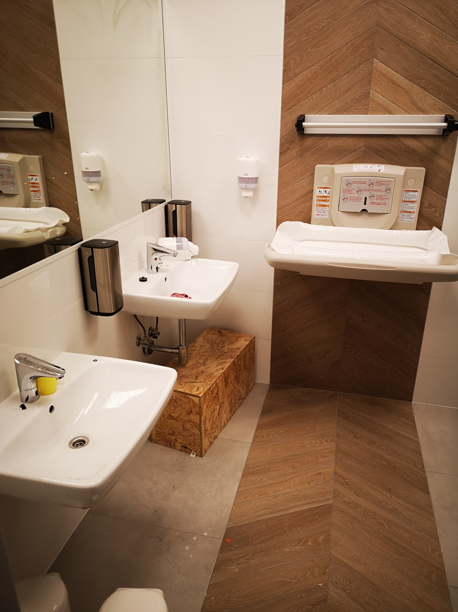 Nowoczesna łazienka publiczna z dwoma umywalkami, dozownikami mydła w metalowych obudowach i przewijakiem dla niemowląt na ścianie z drewnopodobnymi panelami ułożonymi w jodełkę.