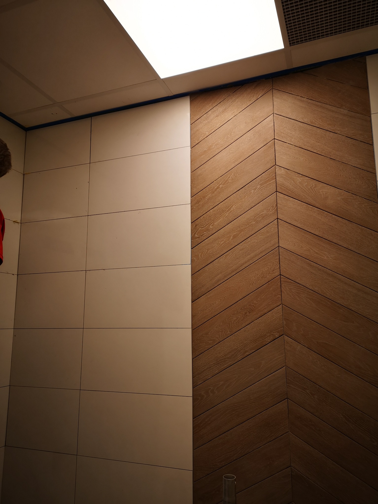 Ściana wykończona jasnymi, kwadratowymi płytkami ceramicznymi po lewej stronie i drewnianą okładziną ułożoną w jodełkę po prawej, oddzielone niebieską taśmą malarską, widoczne oświetlenie sufitowe.