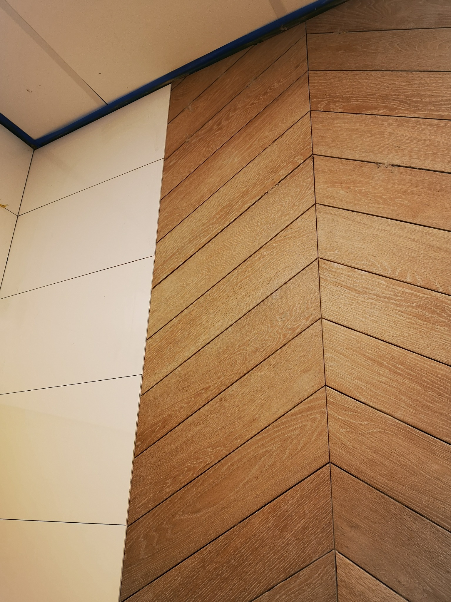 Fragment ściany z ułożonymi w jodełkę drewnopodobnymi płytkami, obok fragment ściany z białymi, prostokątnymi płytkami ceramicznymi, zabezpieczony niebieską taśmą malarską.