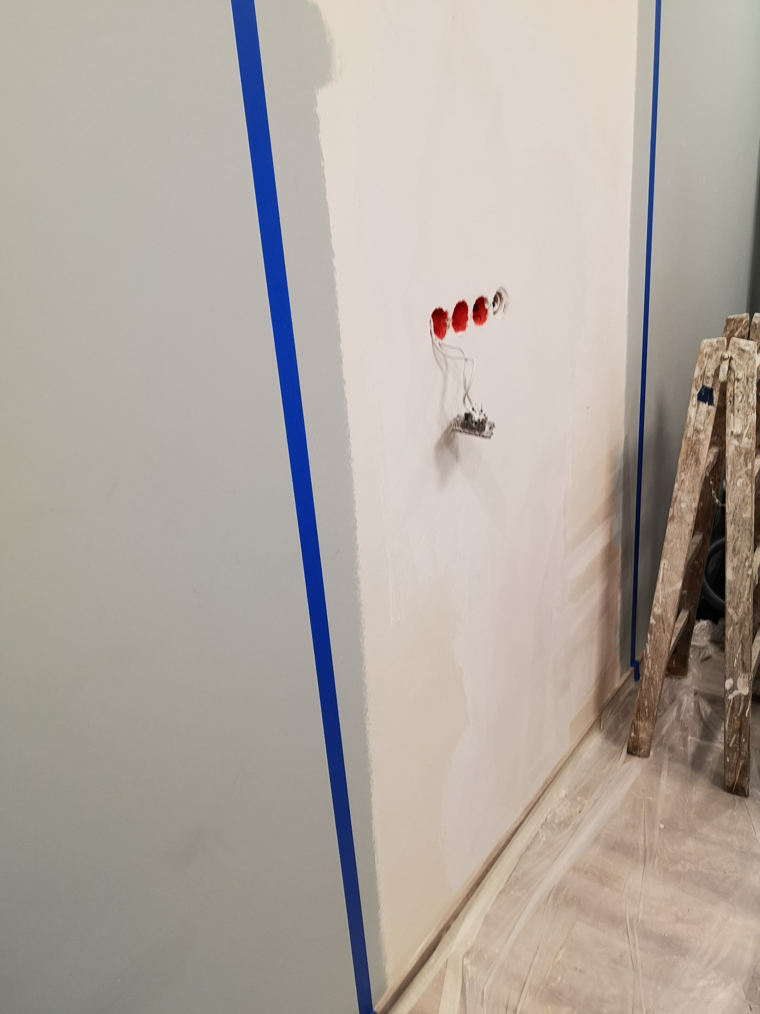 Przygotowanie ściany do malowania: świeżo nałożona gładź, widoczne puszki elektryczne, zabezpieczone taśmą malarską krawędzie i rozłożona folia ochronna na podłodze, drewniana drabina oparta o ścianę.