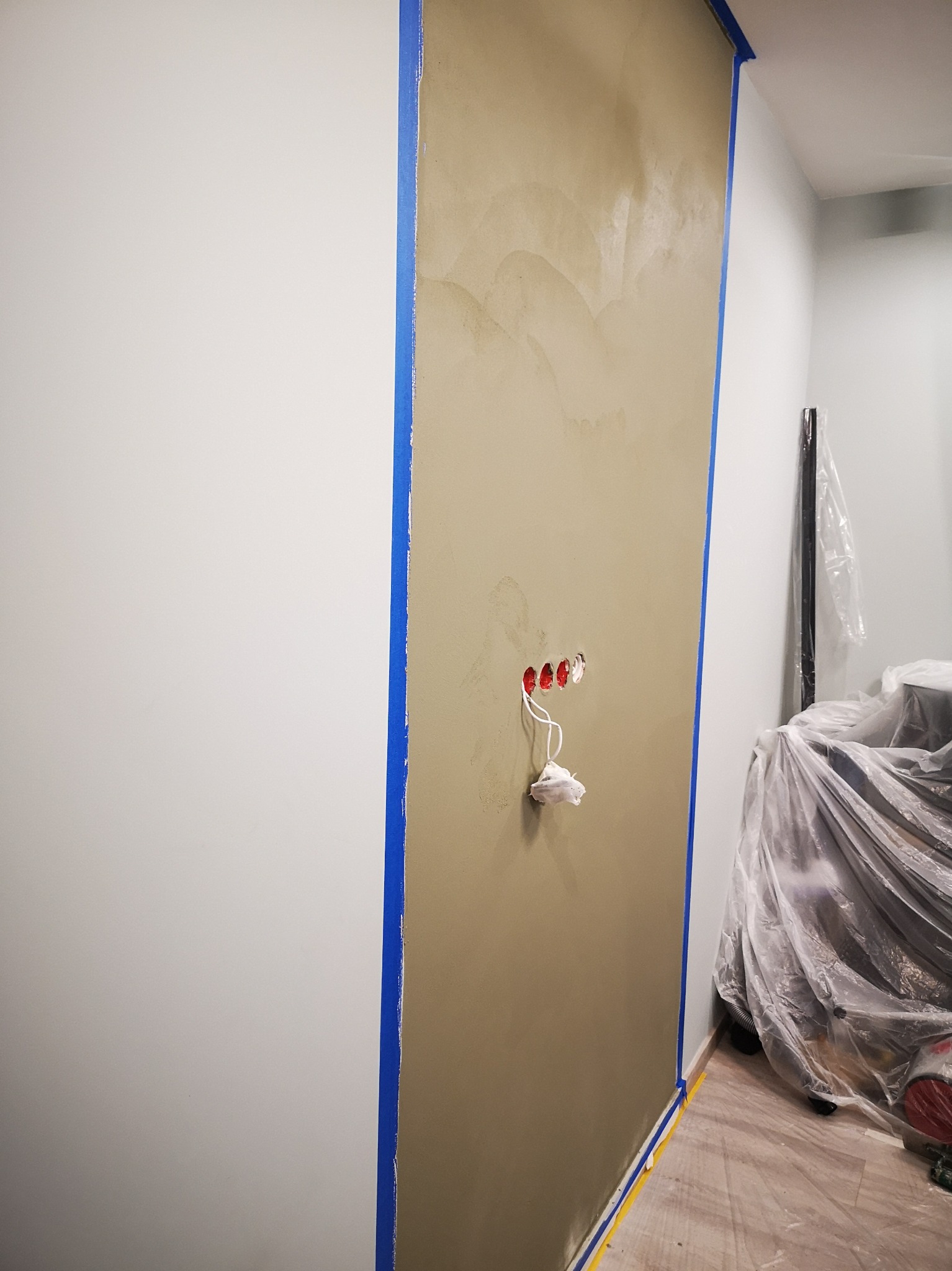 Ściana w trakcie nakładania gładzi z widocznymi puszkami elektrycznymi zabezpieczonymi taśmą malarską, oraz zabezpieczone folią meble w tle.