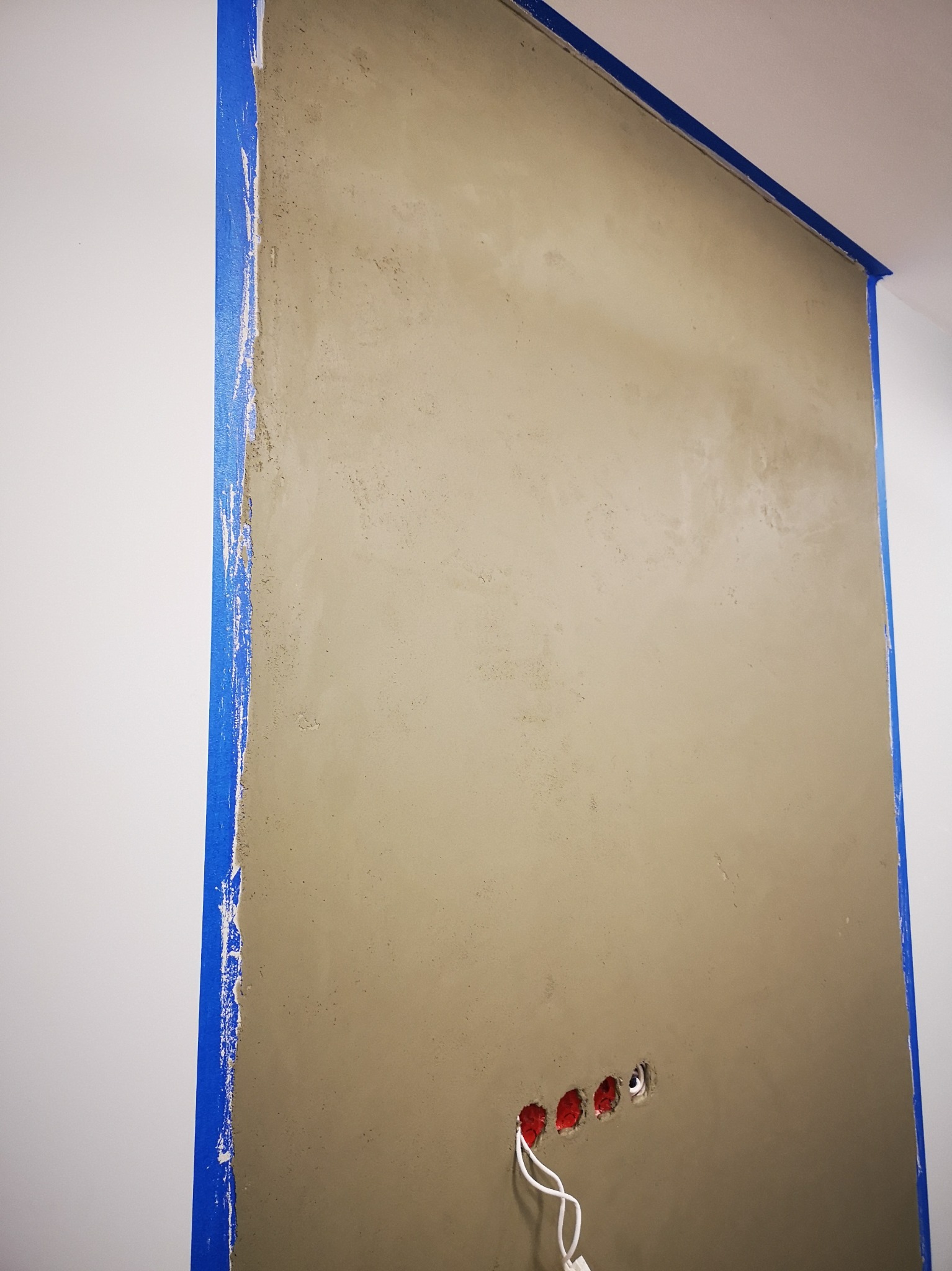 Ściana pokryta świeżą, szarobeżową gładzią, zabezpieczona niebieską taśmą malarską przy białym suficie i ścianie bocznej, z widocznymi otworami na gniazdka elektryczne i wystającymi przewodami.