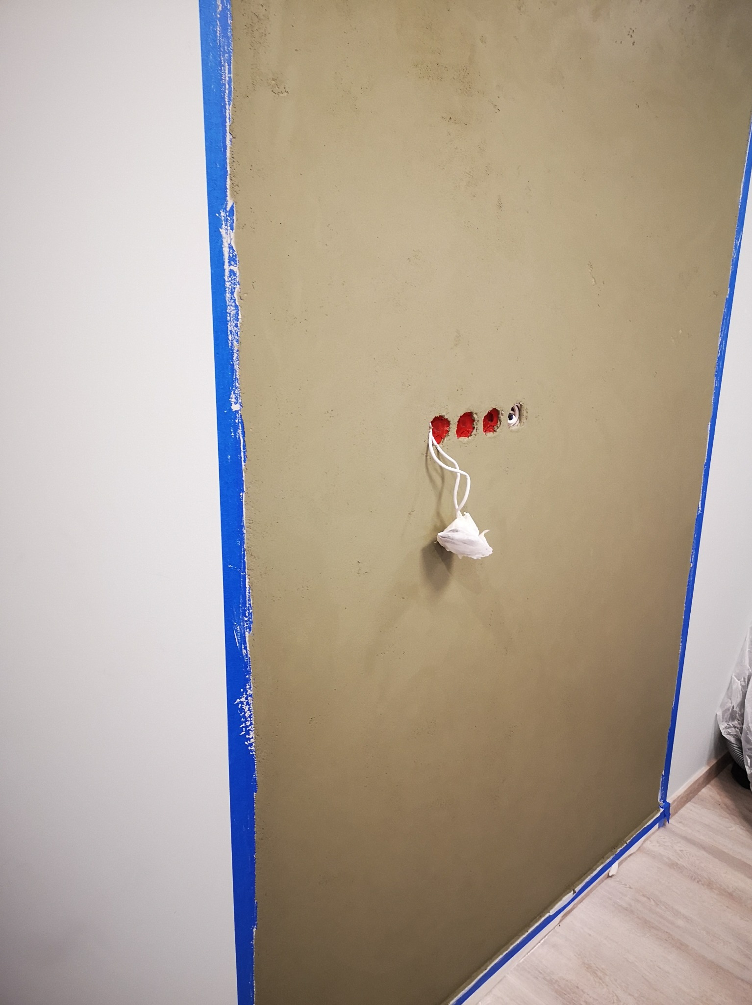 Ściana po nałożeniu gładzi, zabezpieczona niebieską taśmą malarską, z widocznymi otworami na gniazdka elektryczne i wystającymi przewodami zabezpieczonymi folią.