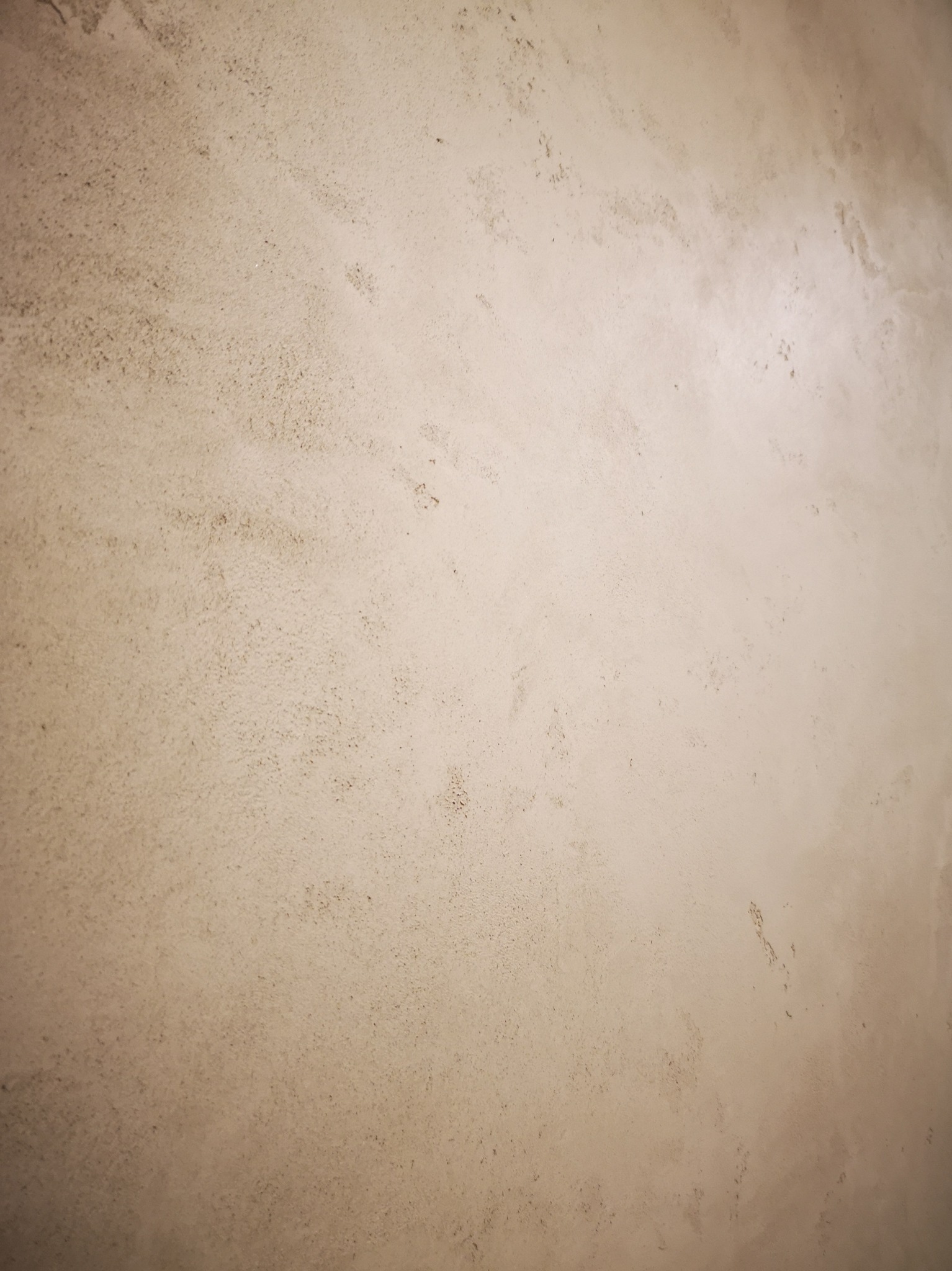 Beżowa ściana z delikatną, nieregularną fakturą przypominającą marmurkowy wzór, oświetlona z góry, ukazująca subtelne cieniowanie i drobne nierówności powierzchni.