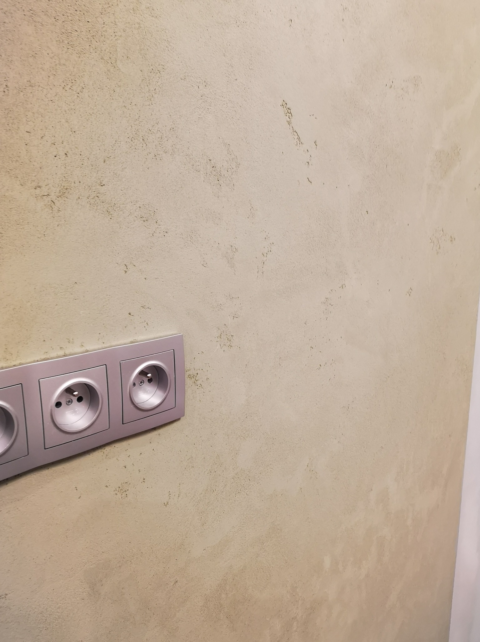 Ściana wykończona w odcieniach beżu z fakturą, listwa z trzema gniazdkami elektrycznymi w kolorze srebrnym.